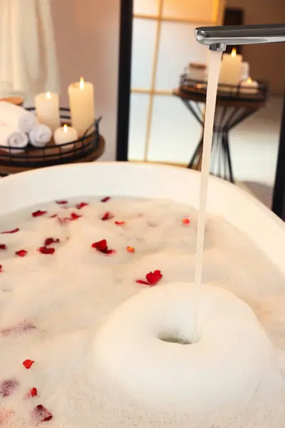 Water pouring into bath tub with foam and red rose petals in bathroom