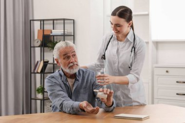 Genç sağlık çalışanı, içerideki ahşap masada haplı ihtiyara bir bardak su veriyor.