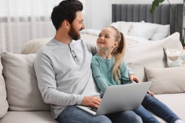 Evdeki kanepede dizüstü bilgisayarlı mutlu bir adam ve kızı var.