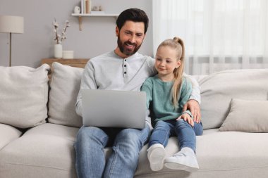 Evdeki kanepede dizüstü bilgisayarlı mutlu bir adam ve kızı var.