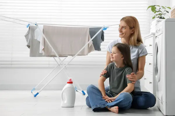 Mother and daughter sitting on floor near fabric softener and clothes dryer in bathroom, space for text