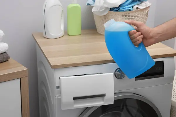 Man with bottle of fabric softener near washing machine indoors, closeup