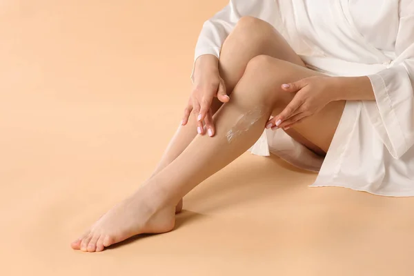 Woman applying body cream onto her smooth legs on beige background, closeup