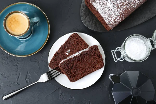 Tasty chocolate sponge cake with powdered sugar and coffee on black textured table, flat lay