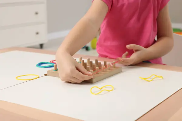 Motor skills development. Girl playing with geoboard and rubber bands at white table indoors, closeup
