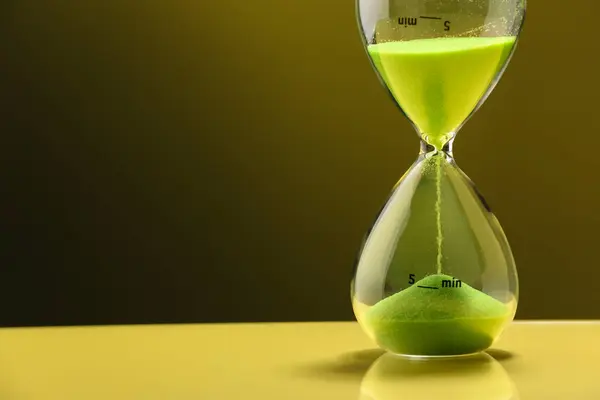 Hourglass with light green flowing sand on table against color background. Space for text