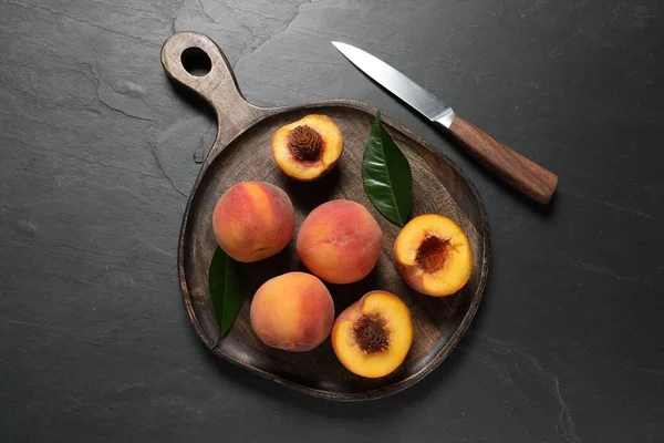 Delicious juicy peaches, leaves and knife on black textured table, top view