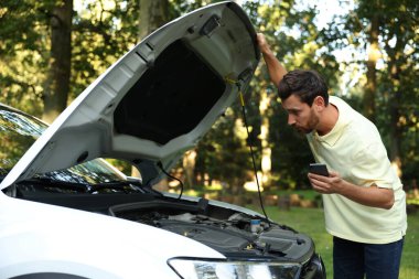 Dışarıda bozuk bir arabanın yanında akıllı telefonu olan üzgün bir adam.