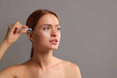 Gri arka planda yüzüne kozmetik serumu uygulayan güzel genç bir kadın.