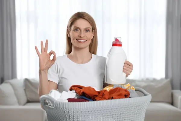 Woman holding fabric softener near basket with dirty clothes and showing OK gesture in room