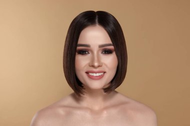 Kahverengi saçlı, bej arka planda gülümseyen güzel bir genç kadının portresi.