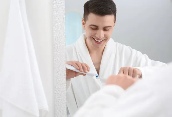 Man squeezing toothpaste from tube onto electric toothbrush in bathroom