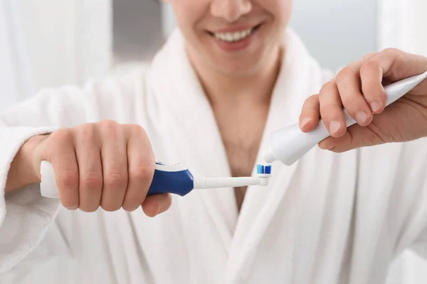 Man squeezing toothpaste from tube onto electric toothbrush indoors, closeup