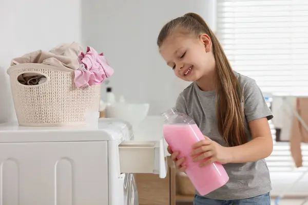 Little girl pouring fabric softener into washing machine in bathroom