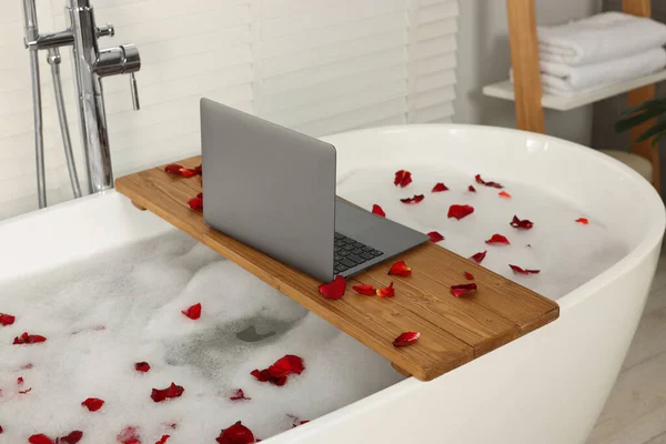 Wooden board with laptop and rose petals on bath tub