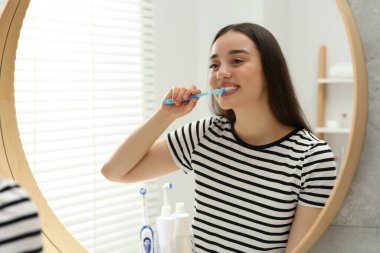 Banyoda aynanın yanında dişlerini plastik diş fırçasıyla fırçalayan genç bir kadın.