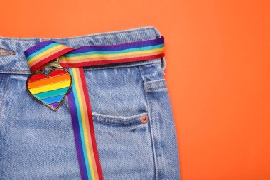 Turuncu arka planda kalp şeklinde kolyesi olan kot pantolon ve gökkuşağı kurdelesi, yazı için üst manzara ve boşluk. LGBT gururu