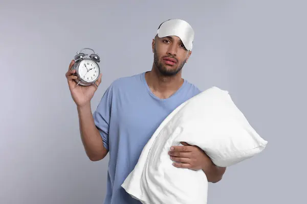 Tired man with pillow, sleep mask and alarm clock on light grey background. Insomnia problem