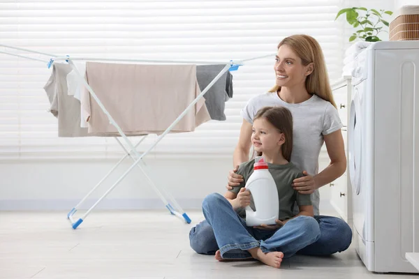 Mother and daughter sitting on floor with fabric softener near clothes dryer in bathroom, space for text