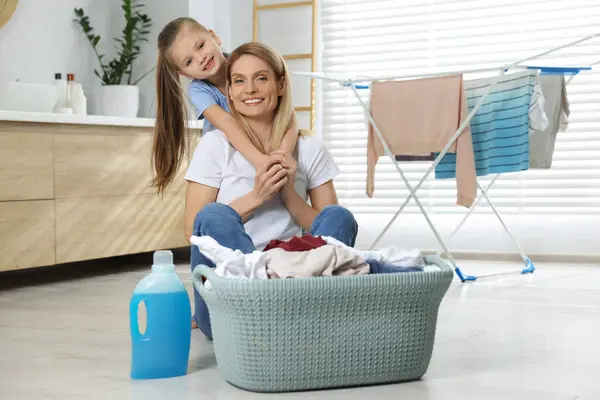 Mother and daughter sitting on floor near basket with dirty clothes, dryer and fabric softener in bathroom