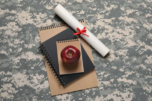 Notebooks, apple and diploma on camouflage background, flat lay. Military education