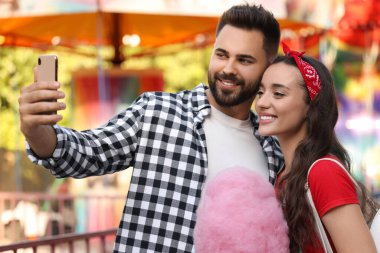 Mutlu genç adam ve kız arkadaşı pamuk şekerle lunaparkta selfie çekiyorlar.