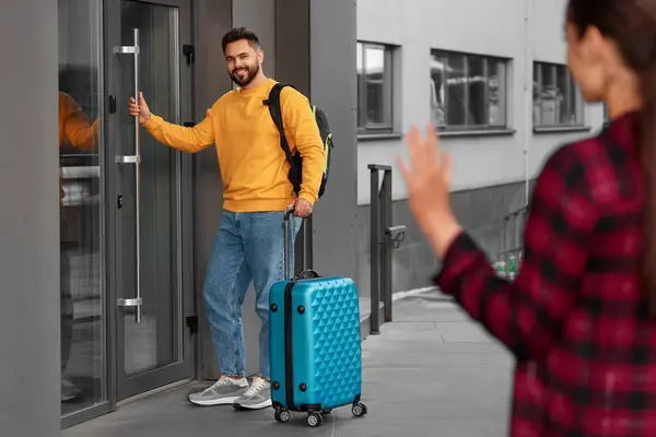 Long-distance relationship. Woman waving to her boyfriend with luggage near building outdoors, selective focus