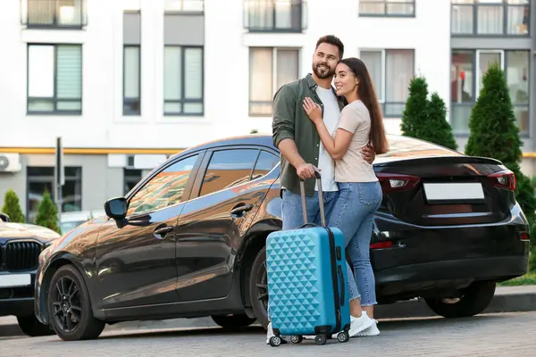 Long-distance relationship. Beautiful young couple with suitcase hugging near car outdoors