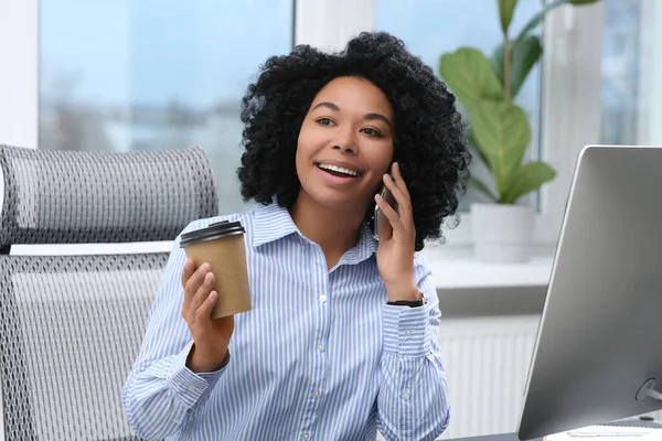 Young woman with cup of drink talking on phone in office