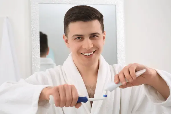 Happy man squeezing toothpaste from tube onto electric toothbrush in bathroom