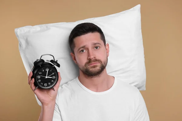 Sleepy man with pillow and alarm clock on beige background. Insomnia problem