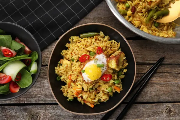 Tasty rice with meat, egg and vegetables in bowl served on wooden table, flat lay