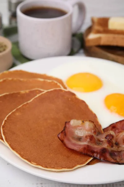 Tasty pancakes with fried eggs and bacon on white wooden table, closeup