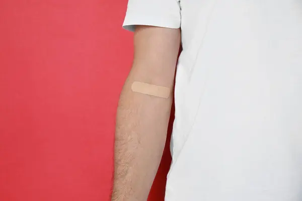 Blood donation concept. Man with adhesive plaster on arm against red background, closeup. Space for text