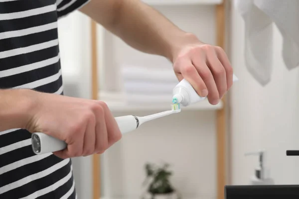 Man squeezing toothpaste from tube onto electric toothbrush in bathroom, closeup