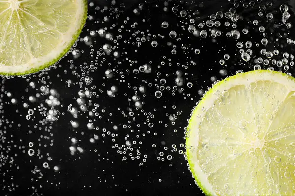 Juicy lime slices in soda water against black background, closeup
