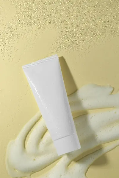 Tube of face cleanser and white foam on beige background, top view. Skin care cosmetic