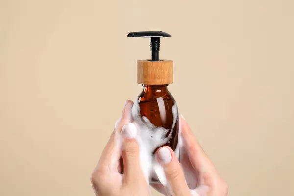 Woman with foam on hands holding bottle of skin cleanser against beige background, closeup. Cosmetic product