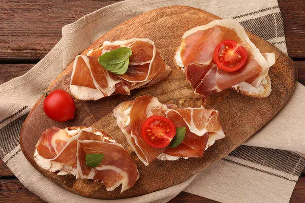 Board of tasty sandwiches with cured ham, basil and tomatoes on wooden table, top view