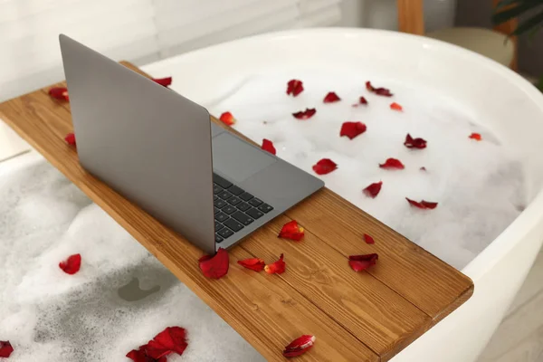 Wooden board with laptop and rose petals on bath tub