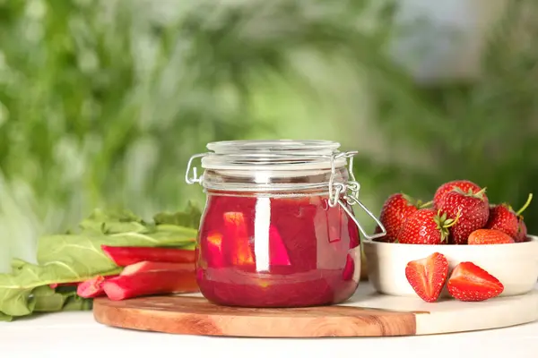 Jar of tasty rhubarb jam, fresh stems and strawberries on white table against blurred background. Space for text