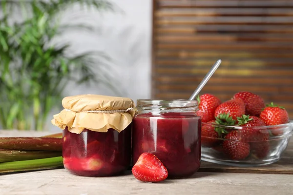 Jars of tasty rhubarb jam, fresh stems and strawberries on wooden table. Space for text