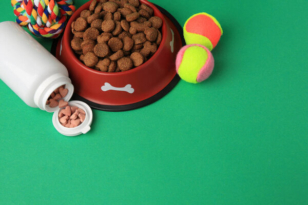 Bowl with dry pet food, bottle of vitamins and toys on green background. Space for text