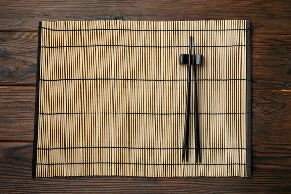 Bamboo mat with pair of black chopsticks and rest on wooden table, top view