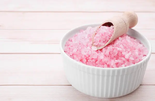 Bowl and scoop with pink sea salt on white wooden table, closeup. Space for text