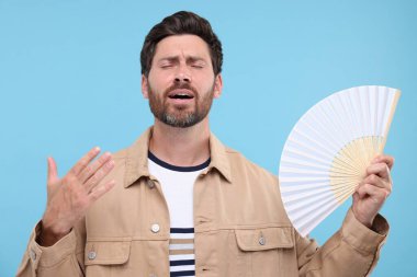 Elinde yelpaze olan mutsuz bir adam açık mavi arka planda sıcaktan muzdarip.