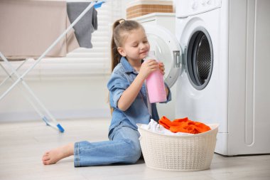 Küçük kız çamaşır makinesi ve sepetin yanında yumuşatıcı kokusu alıyor. Banyoda kirli giysiler, mesaj atmak için yer var.