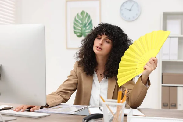Young businesswoman waving hand fan to cool herself at table in office