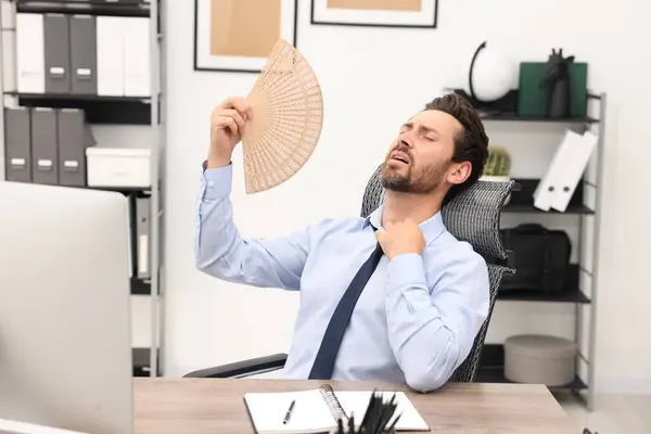 Bearded businessman waving hand fan to cool himself at table in office