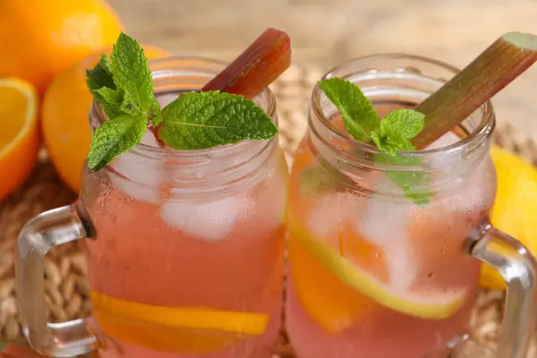 Mason jars of tasty rhubarb cocktail with citrus fruits on table, closeup